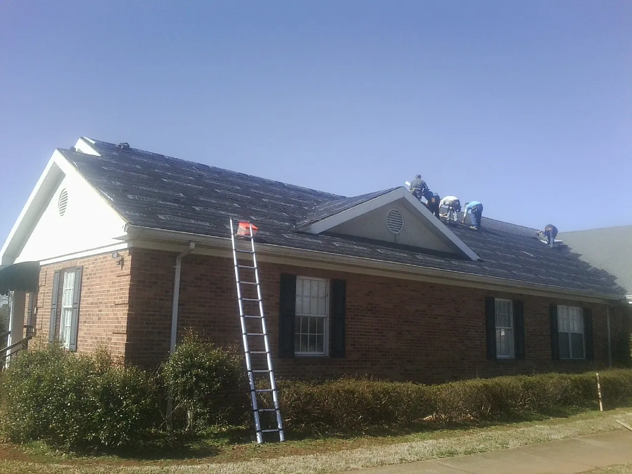 Crew tearing shingles off a brick ranch roof with a ladder set — Emergency Roof Repair work by our crew in Colonial Heights