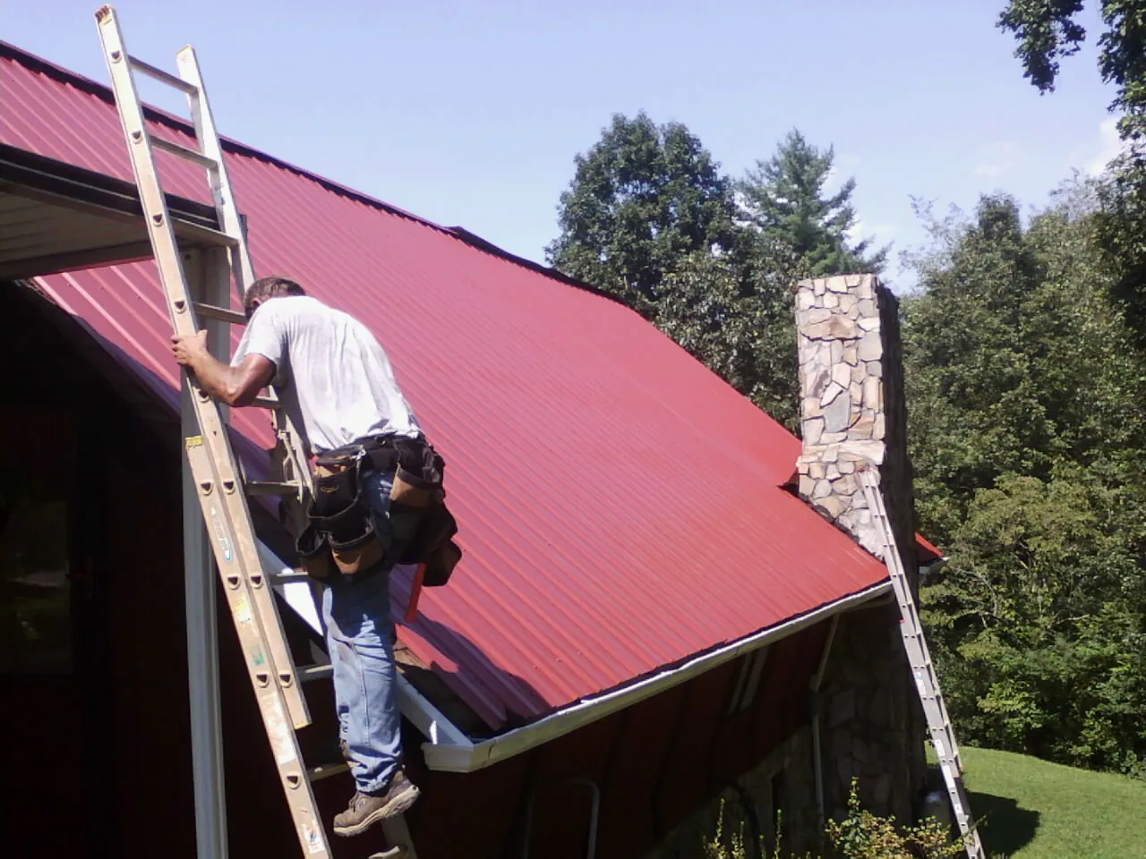 Roofer on a ladder beside a red metal roof and stone chimney — Soffit & Fascia Repair by Roofing Brothers in Colonial Heights